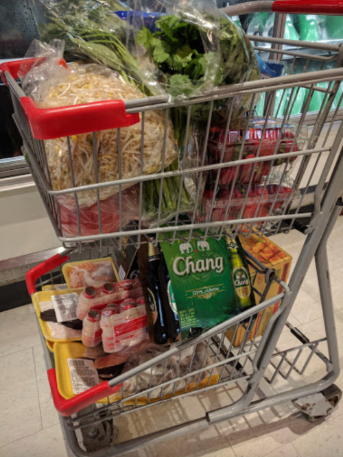 A shopping cart filled with fresh produce, noodles, beer, and packaged meats in a grocery store.